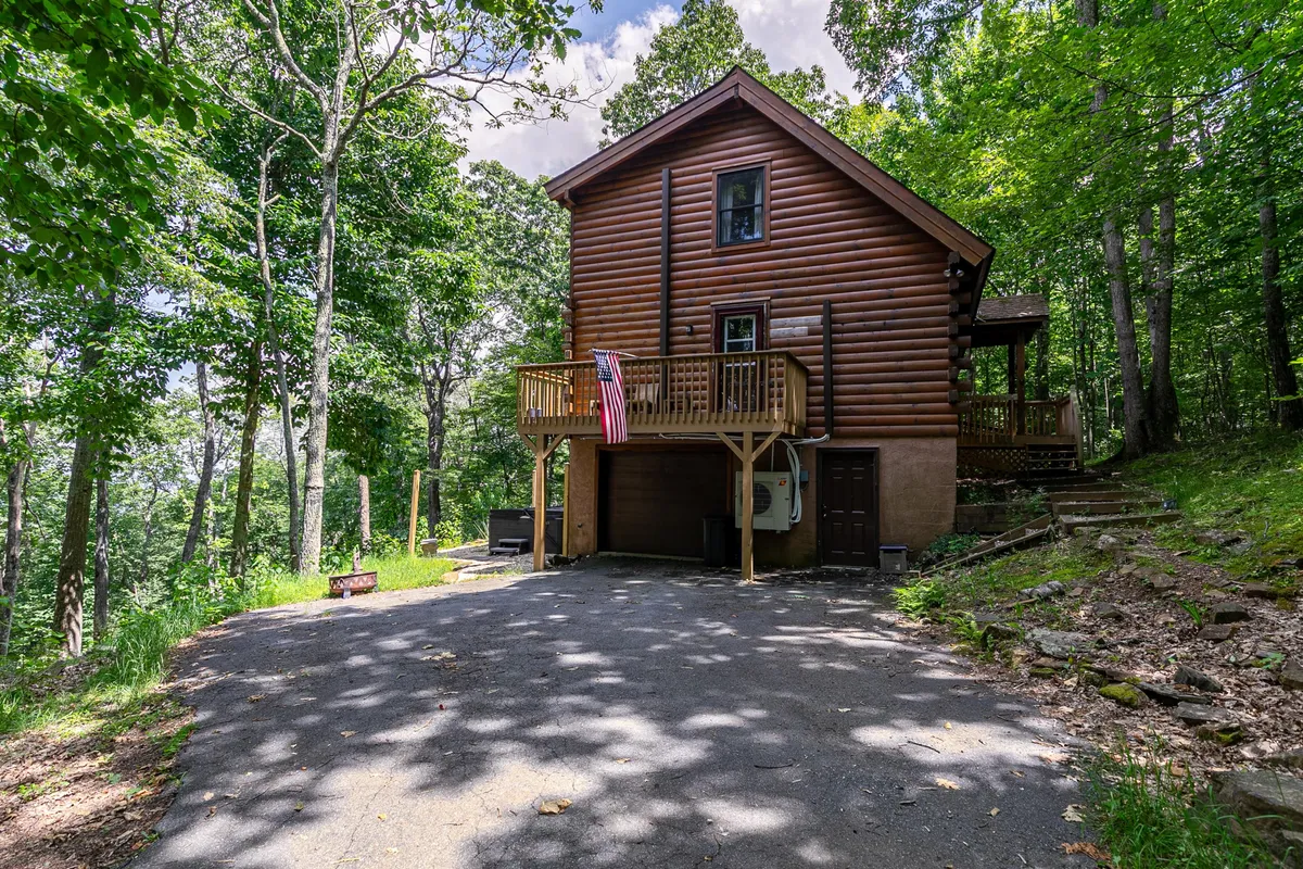 A log cabin nestled in the woods features a spacious deck with a railing and an American flag waving gently in the breeze. The deck offers ample space for relaxing or barbecuing, and the surrounding trees evoke a sense of seclusion and natural beauty. The cabin's rustic exterior enhances the welcoming, pet-friendly atmosphere.