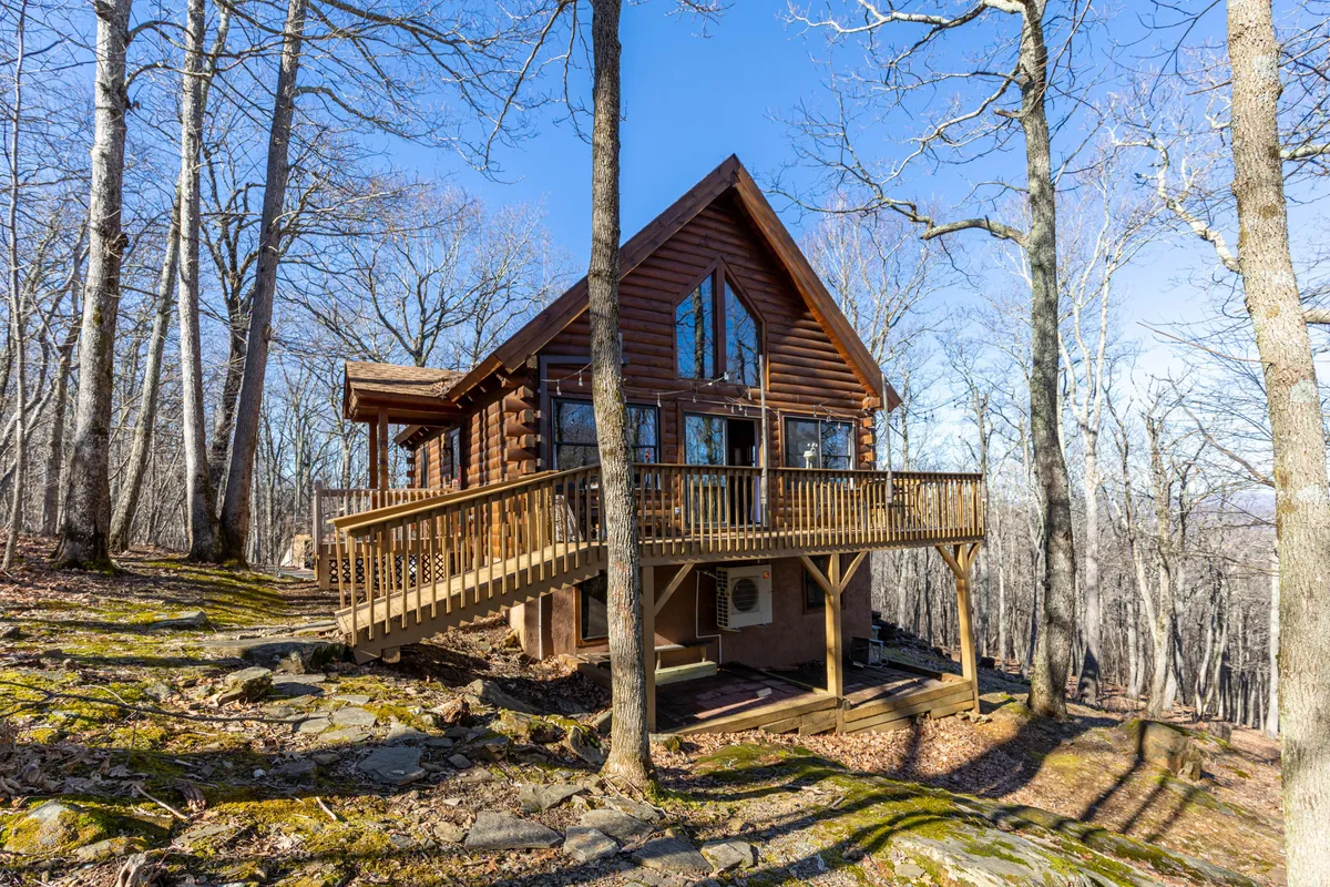 A log cabin with large windows and a spacious deck is nestled among tall trees, blending seamlessly into the surrounding forest. The deck offers a welcoming space for relaxation and socializing, with nature just steps away.