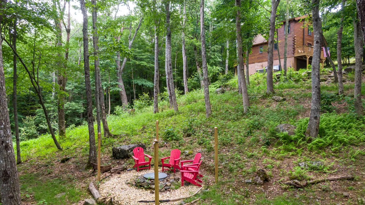 A cozy fire pit area with two red chairs sits surrounded by a wooded landscape, with the cabin in the background. The natural space is perfect for spending time outdoors, enjoying a campfire, and connecting with nature. Ample room around the fire pit hints at space for pets to roam safely.