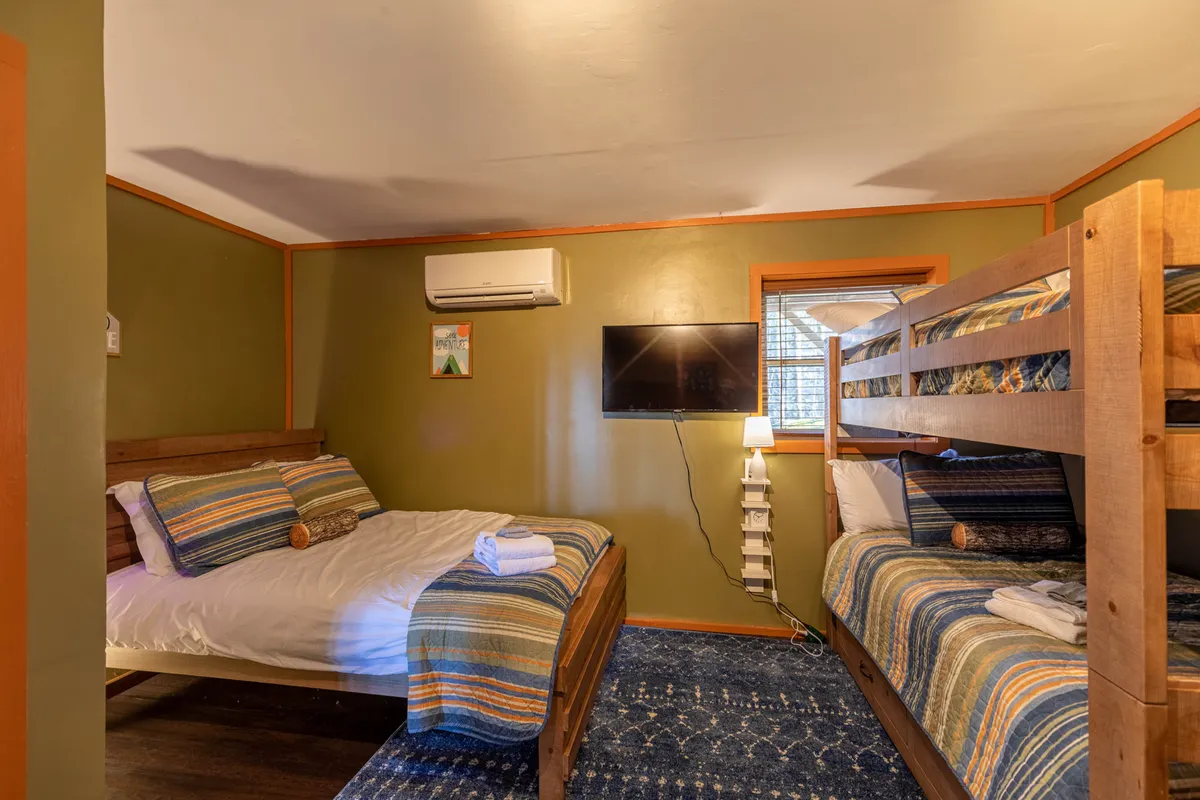 A family-friendly bunk area featuring a twin-over-twin bunk bed, a full-size bed with striped bedding, a television, and a window that brings in daylight. The relaxed arrangement is perfect for kids or friends to unwind, share laughs, or enjoy movie marathons together.
