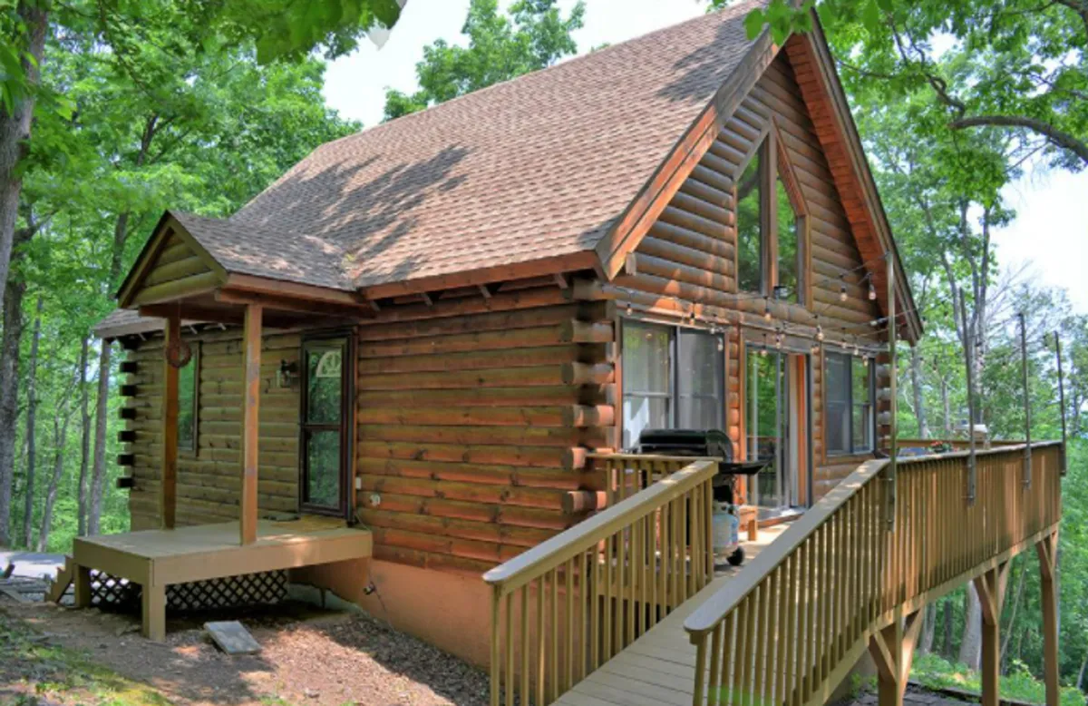 A cozy log cabin set amidst tall trees, with large windows and a spacious deck area visible. The wooden exterior blends into the natural surroundings, and the inviting deck suggests opportunities for outdoor relaxation and barbecuing, while the cabin's placement provides a sense of seclusion and immersion in nature.