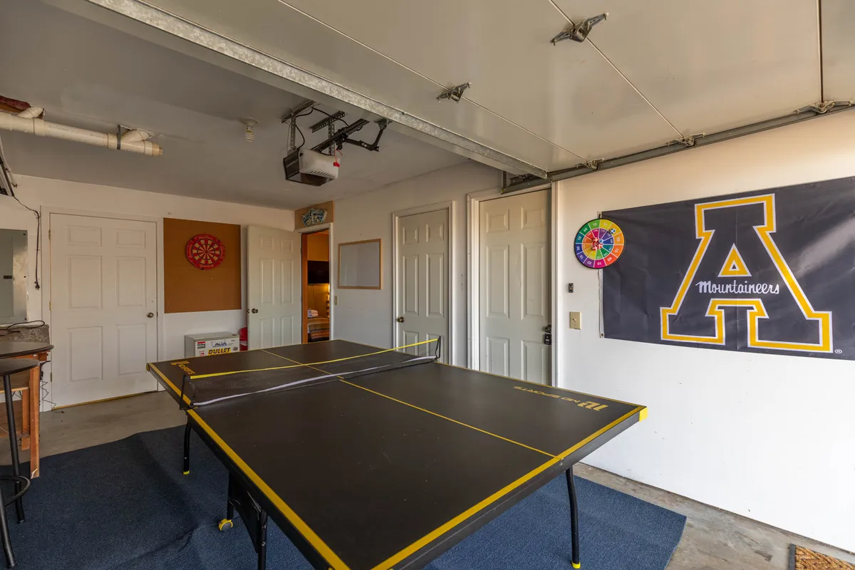 A family-friendly game room with a full-sized ping pong table, dartboard, team banner, and garage doors. The setup encourages friendly competition and group activities, while comfortable seating is visible in the background, creating an energetic yet relaxed environment for making memories.