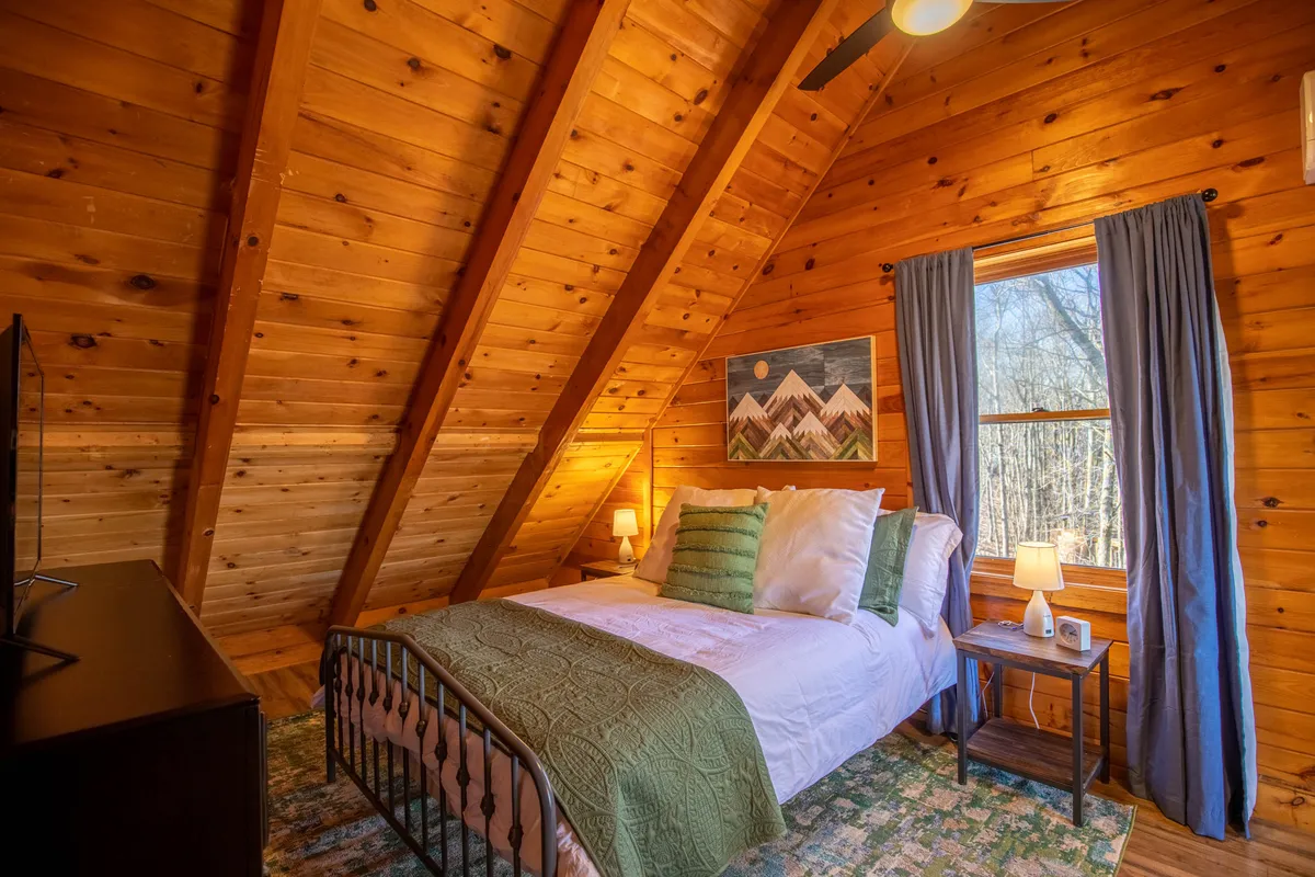 A tranquil bedroom with green and white bedding, wooden walls and ceiling, warm lighting, and tasteful artwork. Nightstands with lamps stand on either side of the bed, and there's a window that lets in gentle natural light, making the space feel calm and private.