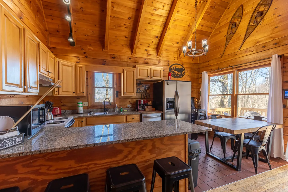 A warm, sunlit kitchen and dining area featuring a granite countertop, wooden cabinets, modern kitchen appliances, and a spacious dining table. Large windows allow in natural light, creating an inviting space ideal for both meal prep and shared gatherings.