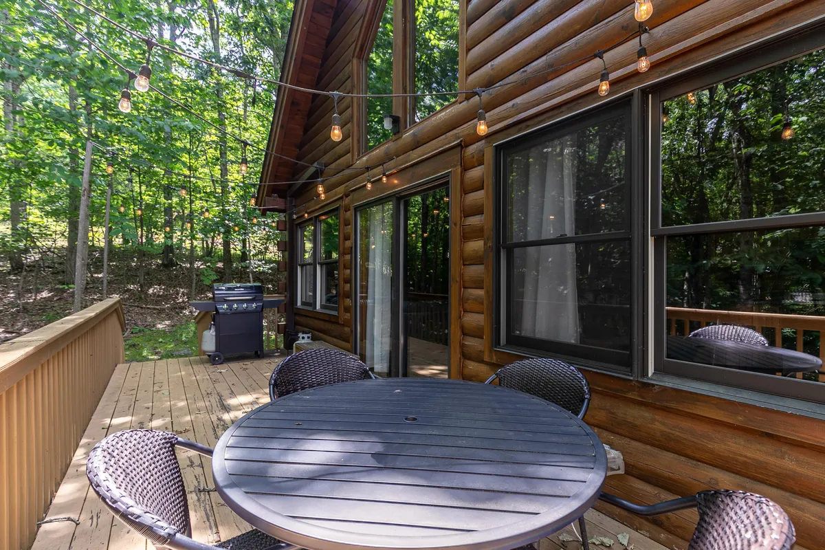A circular table surrounded by woven chairs sits on a patio deck with string lights above, next to the log cabin's exterior. The inviting setup suggests opportunities for outdoor dining or gathering, surrounded by lush greenery and the sounds of nature.