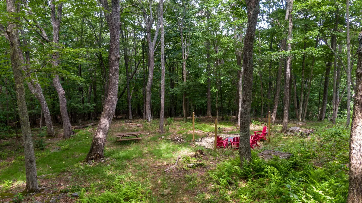 An inviting outdoor firepit area, with red chairs arranged around a campfire, a picnic table nearby, and tall trees enveloping the space. The setting suggests opportunities for campfires, stargazing, and sharing stories in the great outdoors.