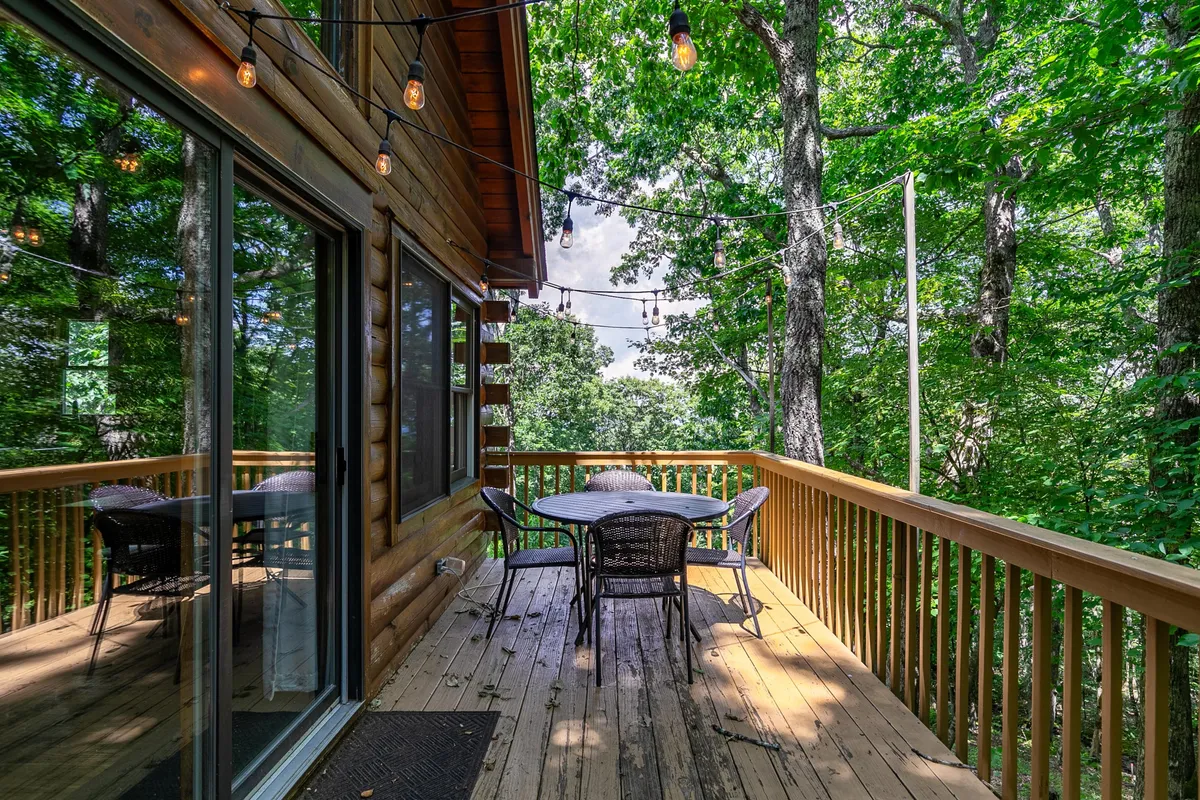 A wooden deck set for outdoor dining beneath a canopy of trees and glowing string lights. The deck is spacious and inviting, ideal for enjoying morning coffee or evening gatherings with pets and friends.