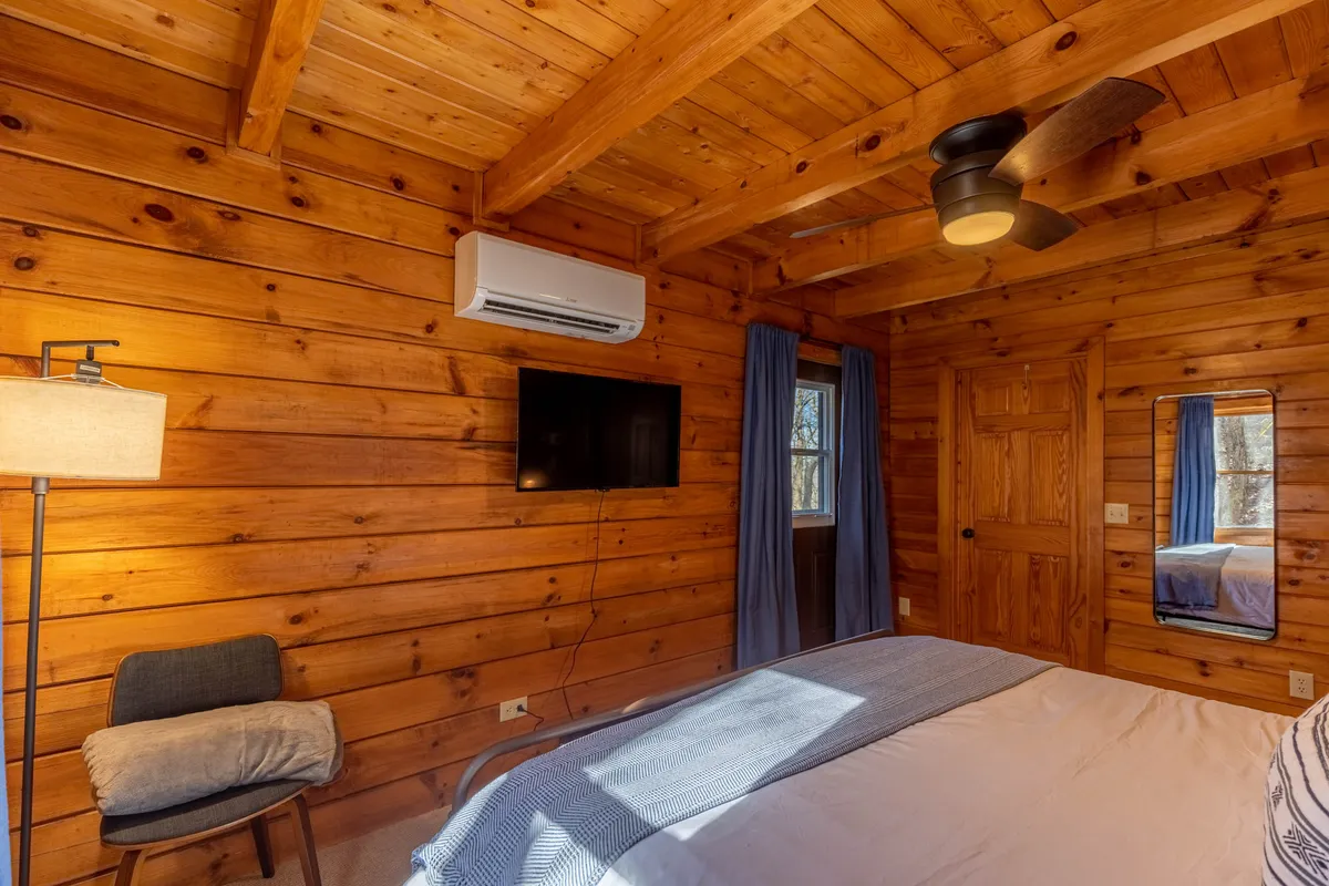 A cozy bedroom with wooden walls, a comfortable bed, ceiling fan, and a television. Warm lamp light fills the space, creating a welcoming and restful atmosphere. Natural light filters in through a window, enhancing the room's tranquil mood.