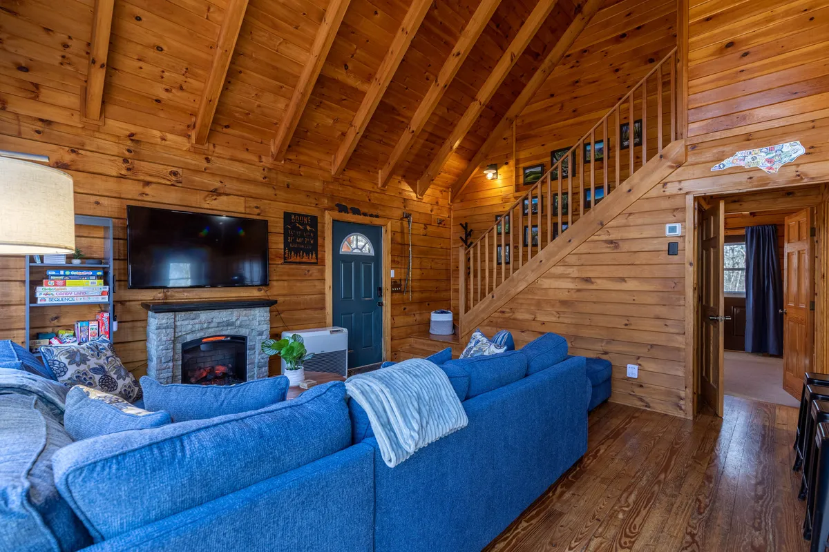 A warm, inviting living room with a plush blue sectional sofa dominates the space, facing a cozy gas log fireplace and a large wall-mounted flat-screen TV. The room features a wooden staircase and decorative wall art, creating a welcoming setting perfect for gathering with family and friends for movie nights or story time.