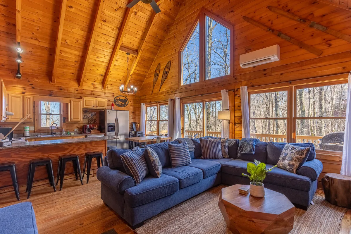 A spacious living area with a large sectional sofa, high wooden-beamed ceiling, and expansive windows that fill the room with sunlight. The scene includes a kitchen island with bar stools, highlighting the seamless connection between living and entertaining spaces, as well as access to the deck with scenic mountain views.