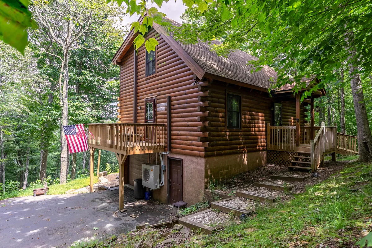 The image shows a charming log cabin nestled on a peaceful mountainside, with a welcoming deck perfect for outdoor relaxation. An American flag flutters beside the inviting structure, surrounded by lush greenery and tranquil natural beauty. The deck hints at opportunities for family gatherings, morning coffee with scenic views, or sunset unwinding, all set in the serene landscape of Boone, North Carolina.
