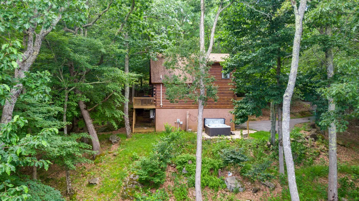 This image captures the log cabin's spacious deck with a hot tub overlooking lush greenery, perfectly illustrating the outdoor relaxation experience. The hot tub sits prominently in the foreground, with the deck's wooden railing and surrounding trees evoking privacy and immersion in nature.