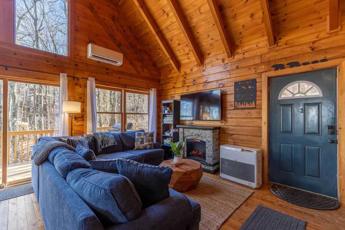 A spacious, warmly lit living room featuring a comfortable sofa, a stone fireplace, large windows that let in daylight, and a television. Seasonal decorations add a welcoming, festive touch, while the layout invites guests to gather, relax, and share stories.