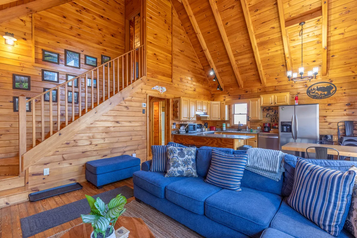 A wide-angle view of the open-concept living, dining, and kitchen area, with a sofa, staircase, and a clear view of both the kitchen and dining table. The scene is staged for family gatherings, game nights, and shared meals, capturing the space’s versatility.