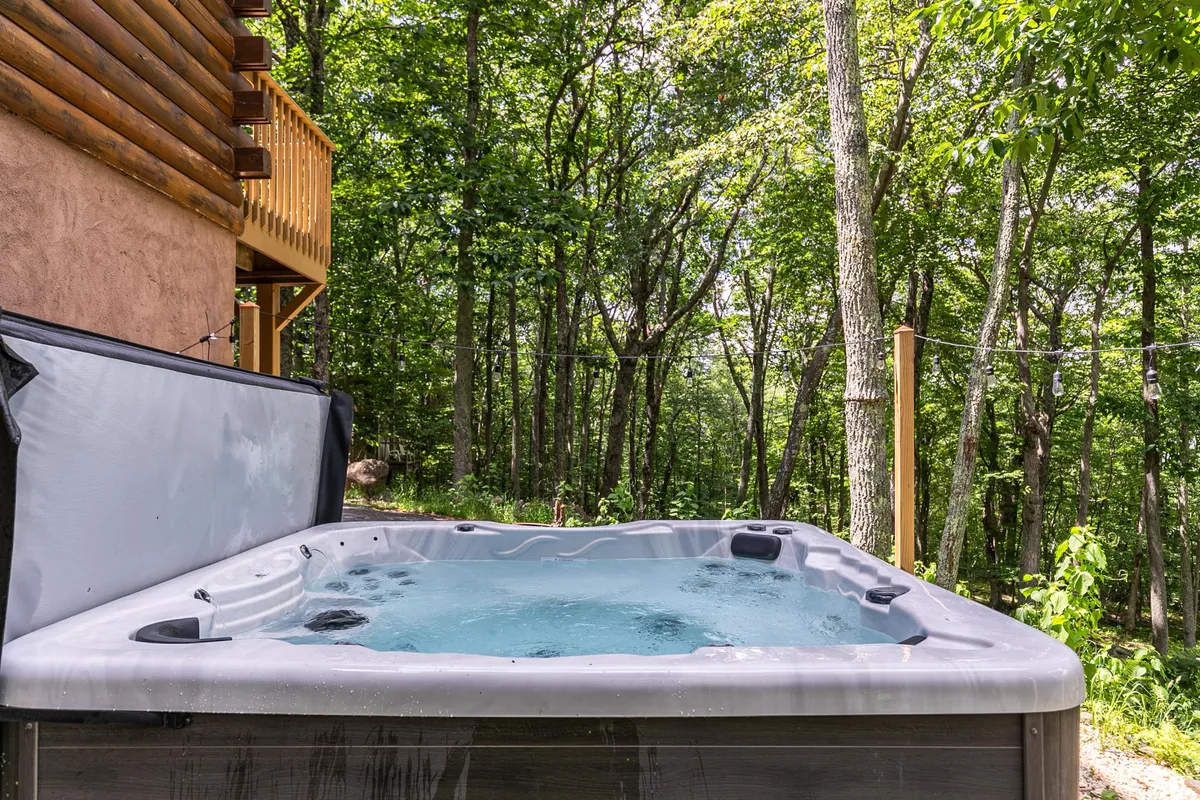 An inviting outdoor hot tub sits on a wooden balcony, surrounded by mature trees. The setting is tranquil, offering views of the forest and an open sky above, suggesting the perfect place to relax and socialize while immersed in nature.