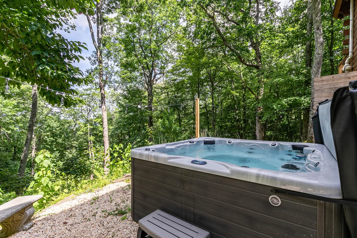 A tranquil outdoor hot tub surrounded by trees and natural landscaping. The gravel pathway leads up to the hot tub, which is set against a backdrop of lush greenery, offering a private retreat for relaxation and nature immersion.